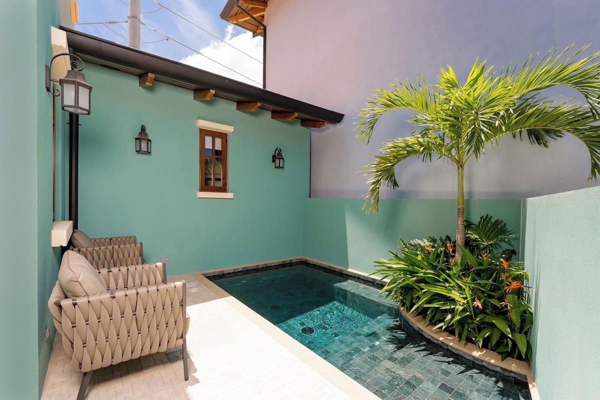Una piscina con deck de azulejos, dos sillas de descanso, una palmera y faroles en la pared.