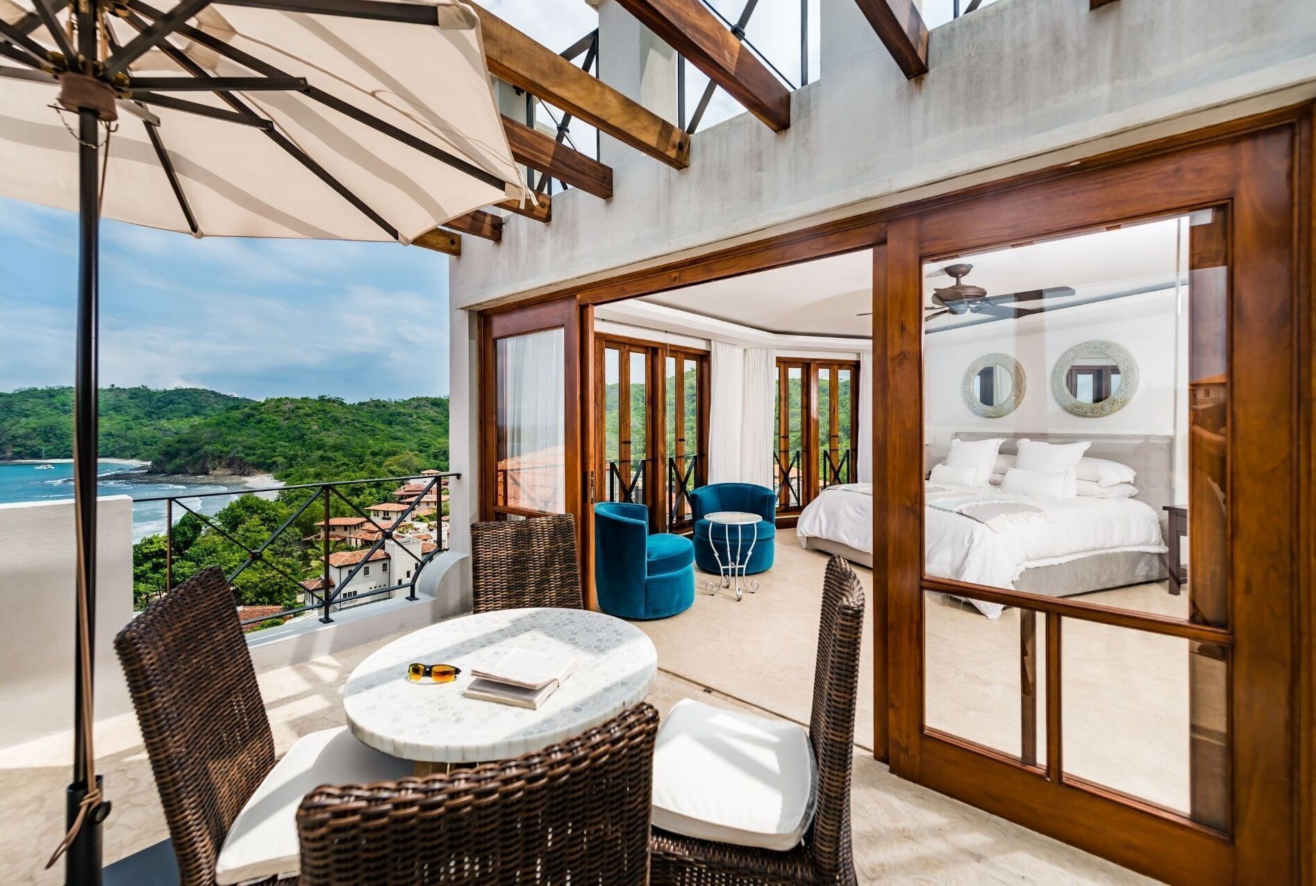 Un balcon agrémenté de meubles en osier, d’une table ronde et offrant une vue sur la mer et la verdure.