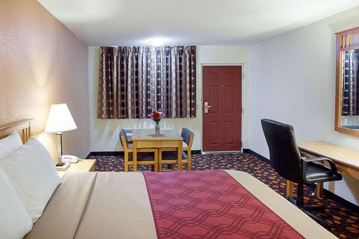 A hotel room with a bed, a desk, a chair, a table with a vase of flowers, and a patterned carpet.