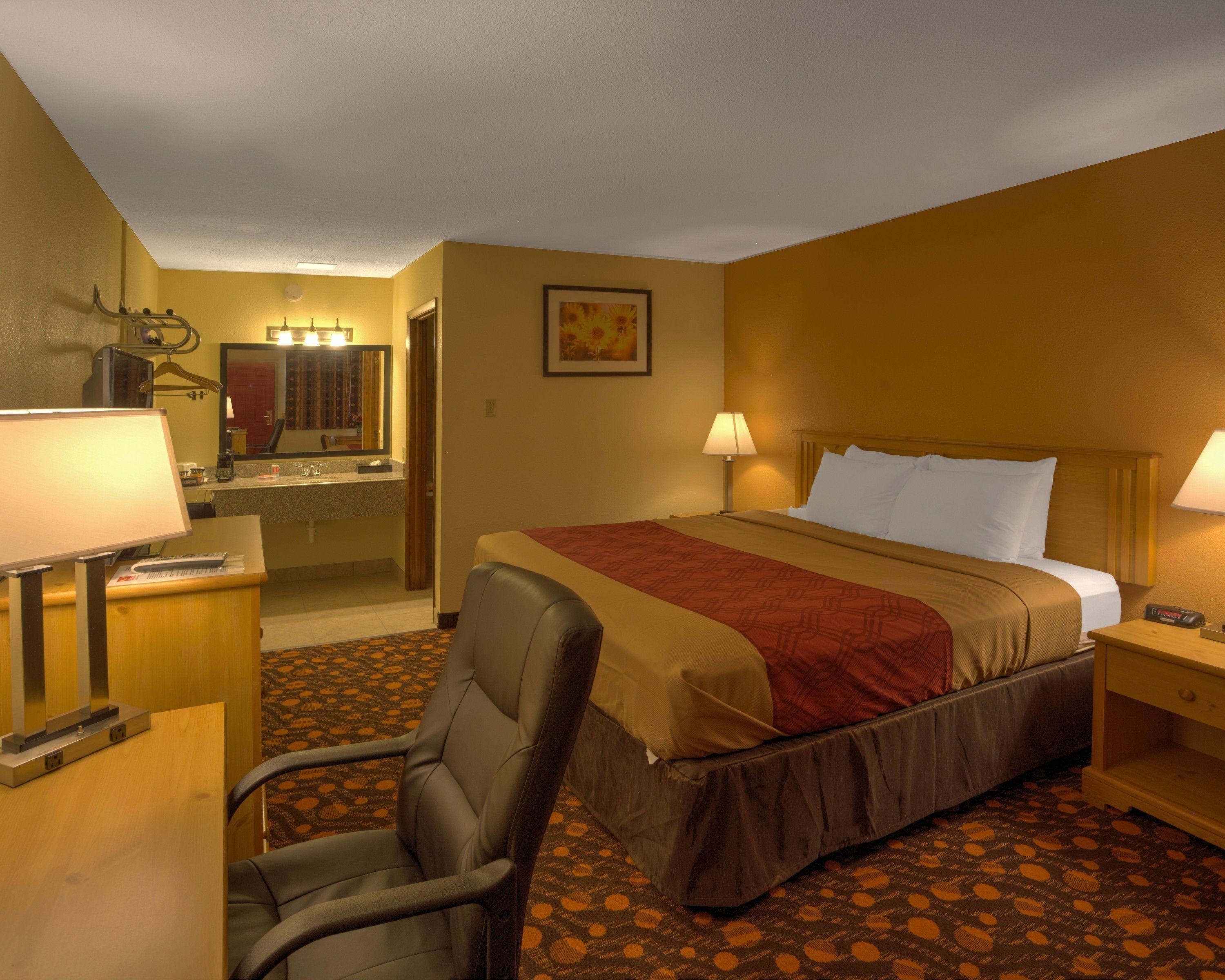 Une chambre d’hôtel comprenant un lit, un bureau avec une lampe, une chaise, une salle de bain avec un miroir et une moquette à motifs.