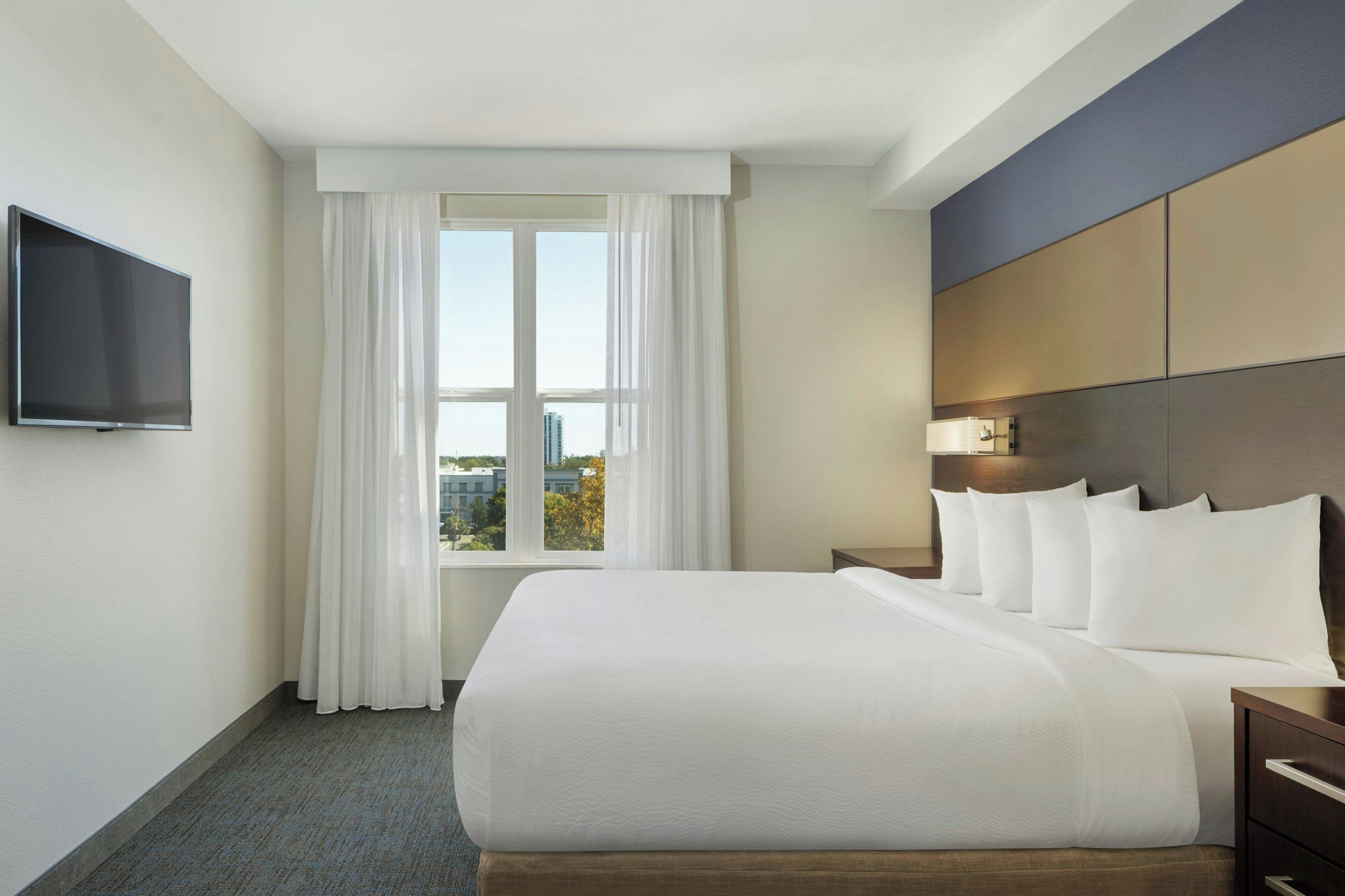 Habitación de hotel con una cama grande, un televisor de pantalla plana montado en la pared, una ventana con cortinas blancas y vistas a edificios.
