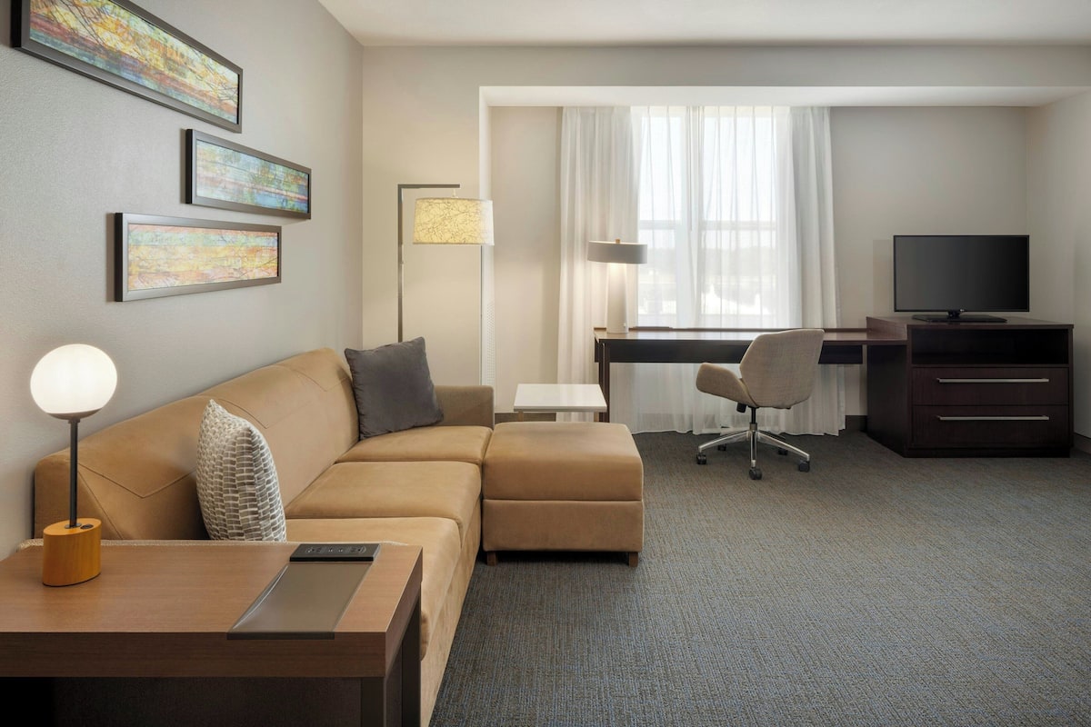 Ein Hotelzimmer mit Sofa, Schreibtisch, Stuhl, Fernseher, Lampe und einem Kunstwerk an der Wand.