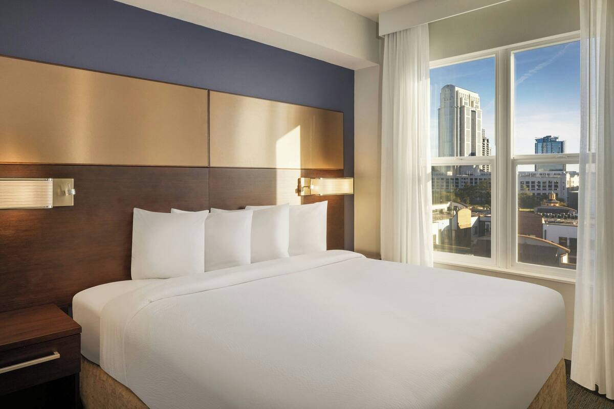 Ein Hotelzimmer mit einem großen Bett, einem Holzkopfteil, einem Nachttisch und Blick auf die Stadt durch große Fenster.