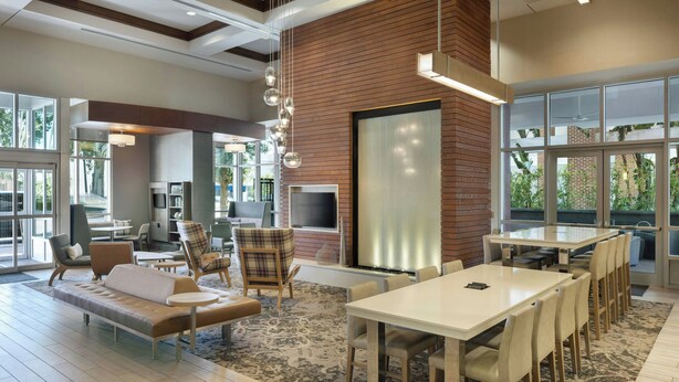 A spacious living area with a large sofa, dining table, and chairs, a fireplace, and large windows offering a view of greenery. - Residence Inn by Marriott Orlando Downtown (Orlando)
