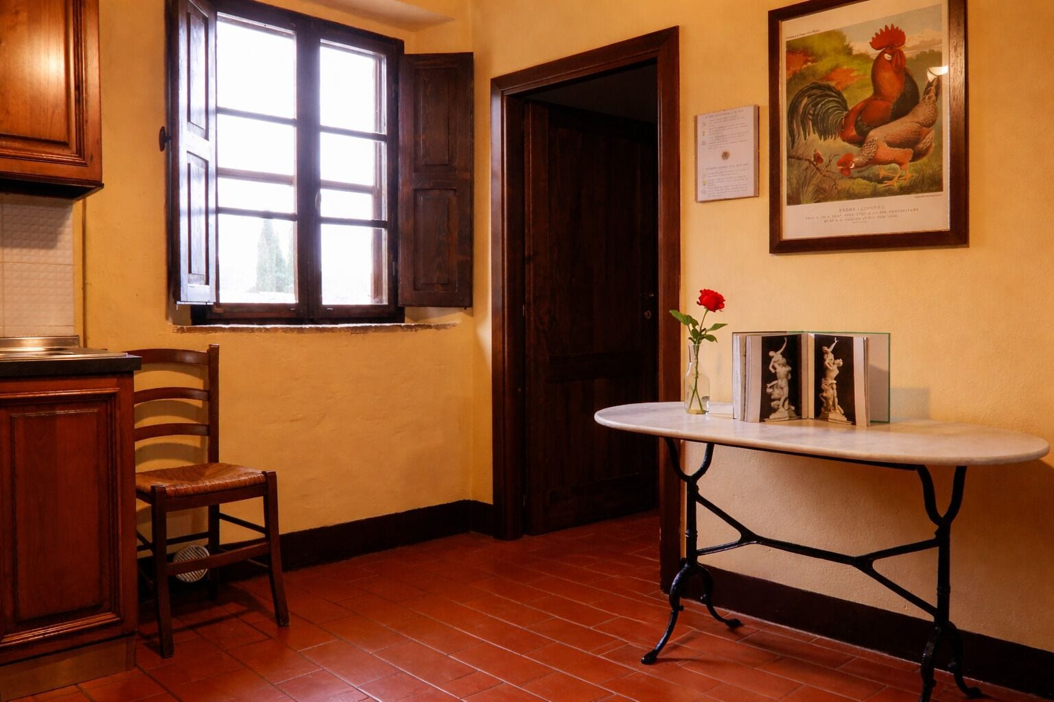 Una cocina con armarios de madera, una ventana con contraventanas, una puerta, una mesa con un florero y libros, y un cuadro de un gallo enmarcado en la pared.
