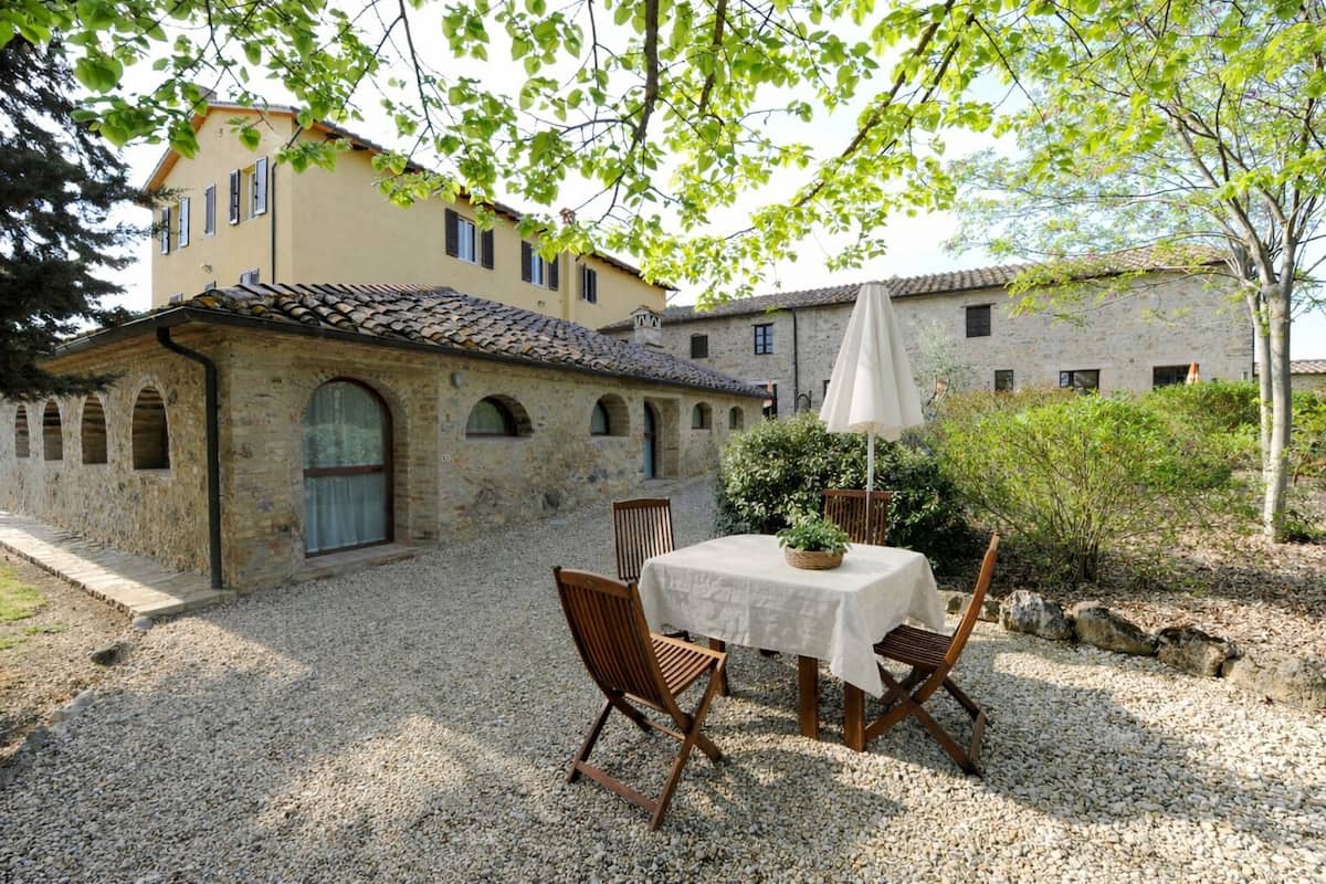 Un patio con una mesa y sillas, un edificio con ventanas de arco y un muro de piedra.