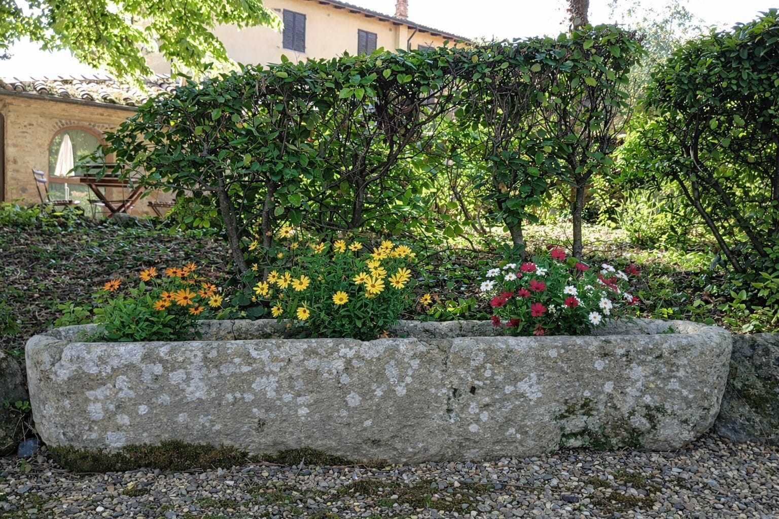 A stone planter with various flowers and a garden area with a building in the background.