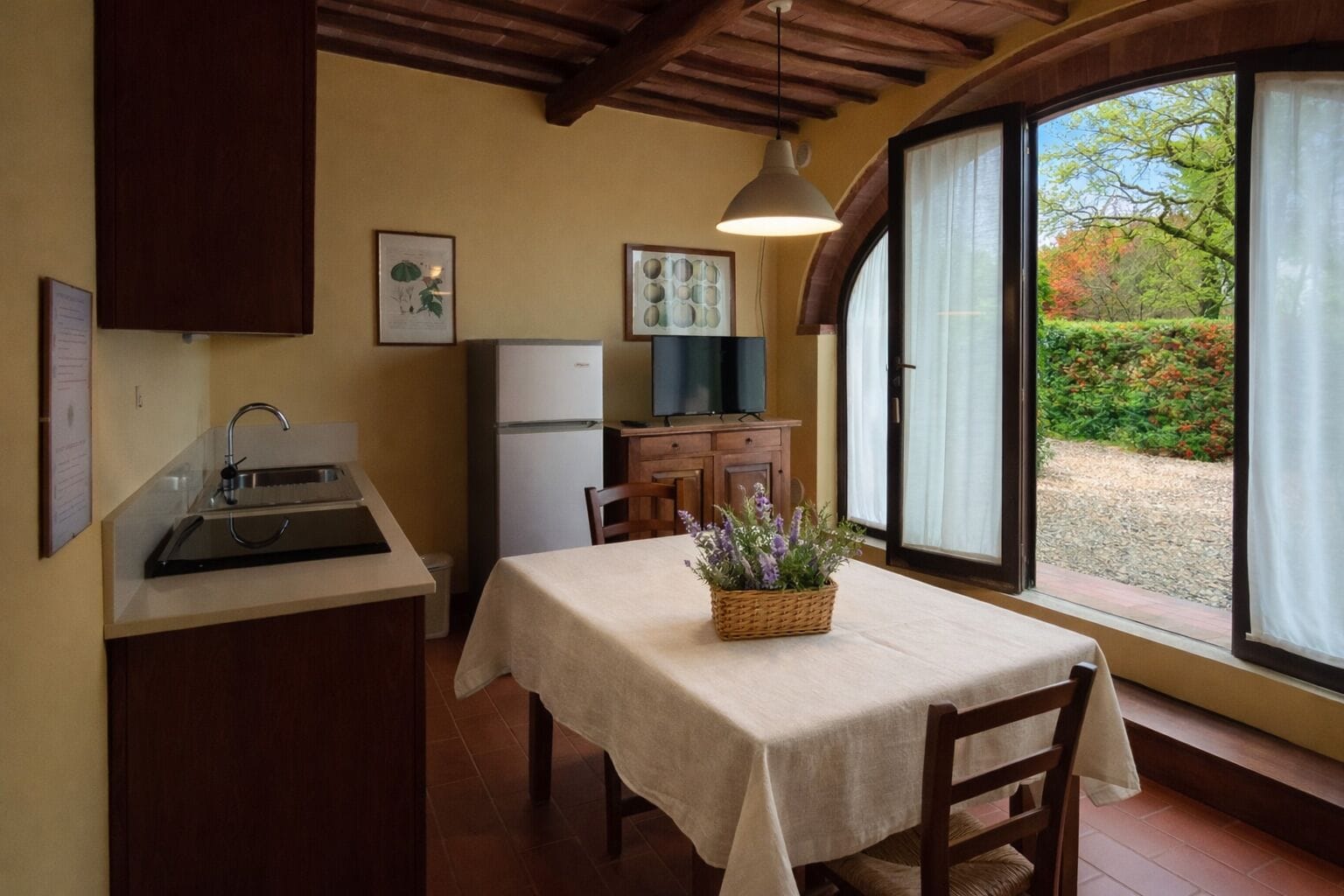 A kitchen with a sink, a refrigerator, a dining table with a white tablecloth, and a window with a view of greenery.