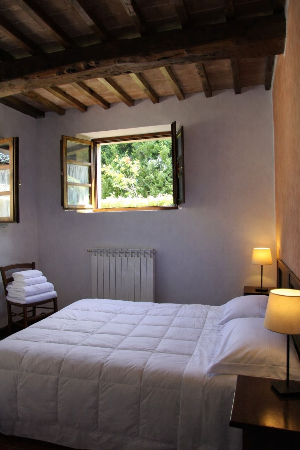 Une chambre avec un lit, une fenêtre donnant sur un espace verdoyant, un radiateur, une chaise avec des serviettes et une table de chevet avec une lampe.