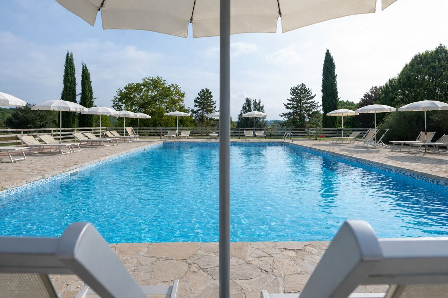 Una piscina con sillas de descanso y sombrillas, rodeada de árboles y una terraza de piedra.