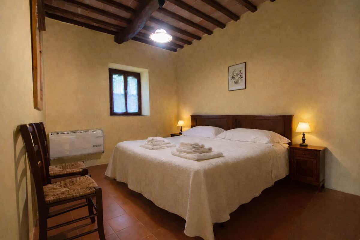 Un dormitorio con una cama doble, dos lámparas de noche, un radiador, una ventana y una silla.