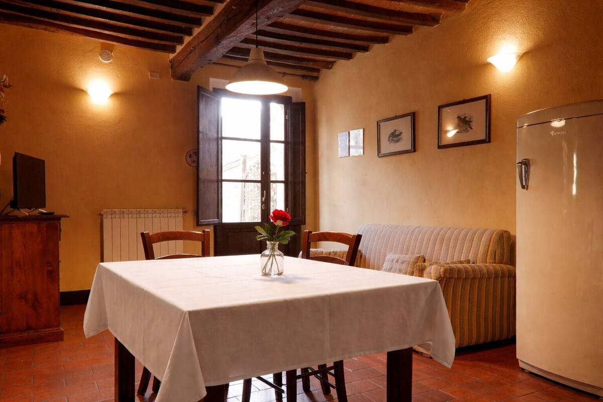 Un comedor con una mesa, sillas, un florero con una flor, un sofá, un radiador y un refrigerador.