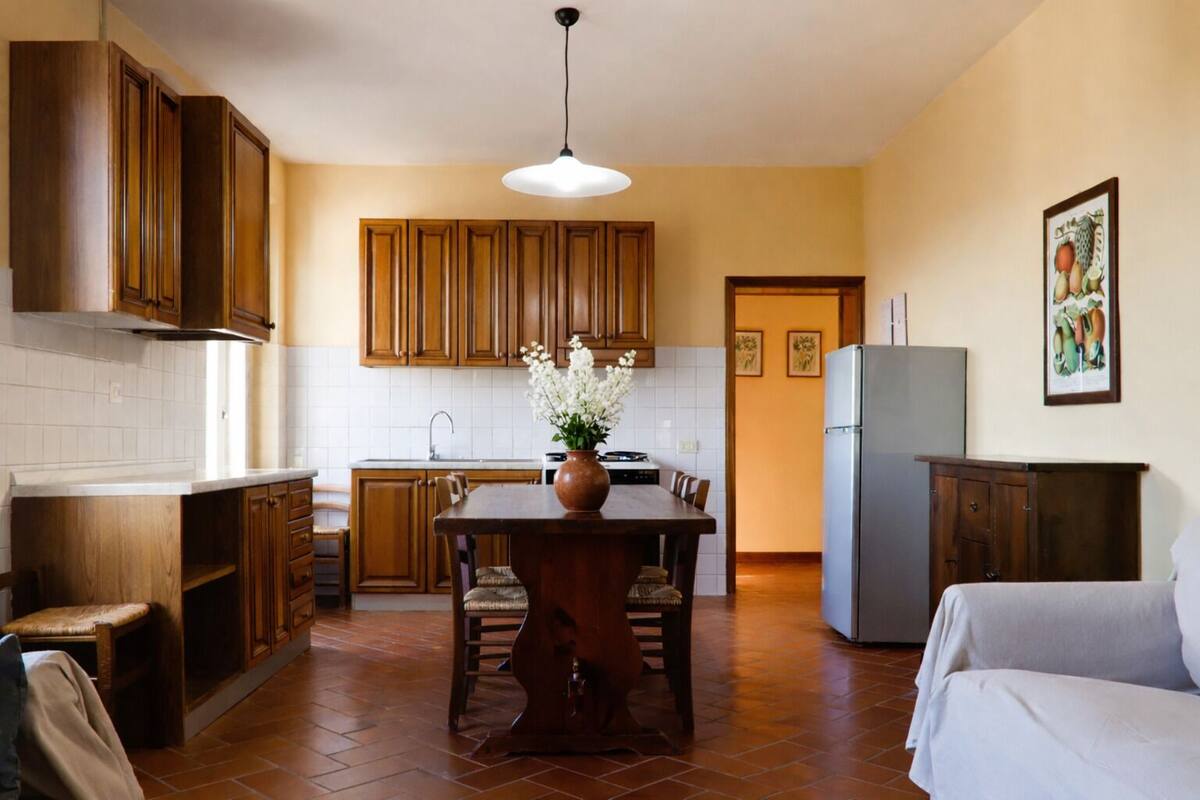 Cocina compacta con armarios de madera, una mesa de comedor con sillas, un refrigerador y una lámpara colgante.
