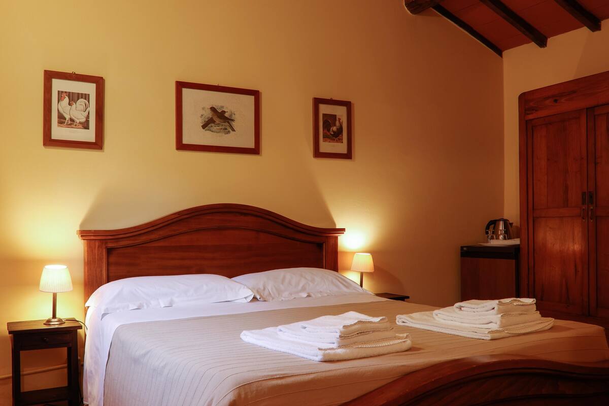 Habitación de hotel con cama de madera, dos cuadros en la pared, una mesita de noche con lámpara y un armario de madera.