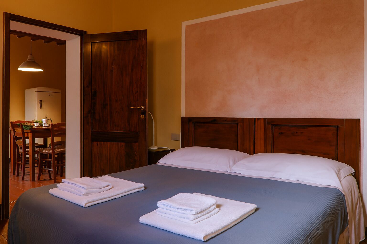 Ein Hotelzimmer mit Bett, zwei Handtüchern, einem hölzernen Kopfteil, einer Holztür und einem Essbereich mit Stühlen und Tisch.