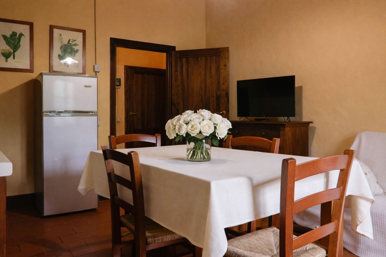 A dining area with a table set for four, a white refrigerator, a wooden cabinet, and a flat-screen TV.