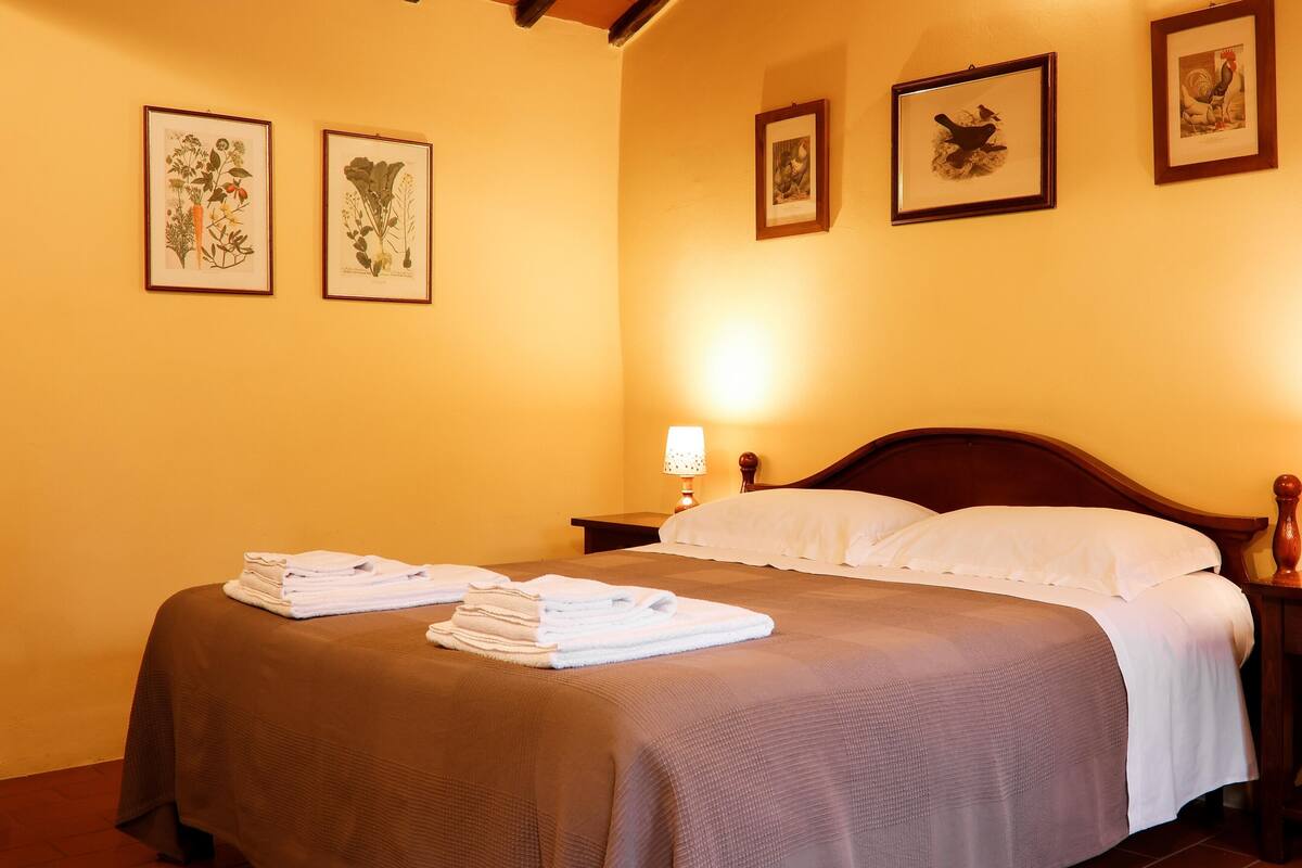 Habitación de hotel con cama, mesita de noche con lámpara y tres láminas botánicas enmarcadas en la pared.
