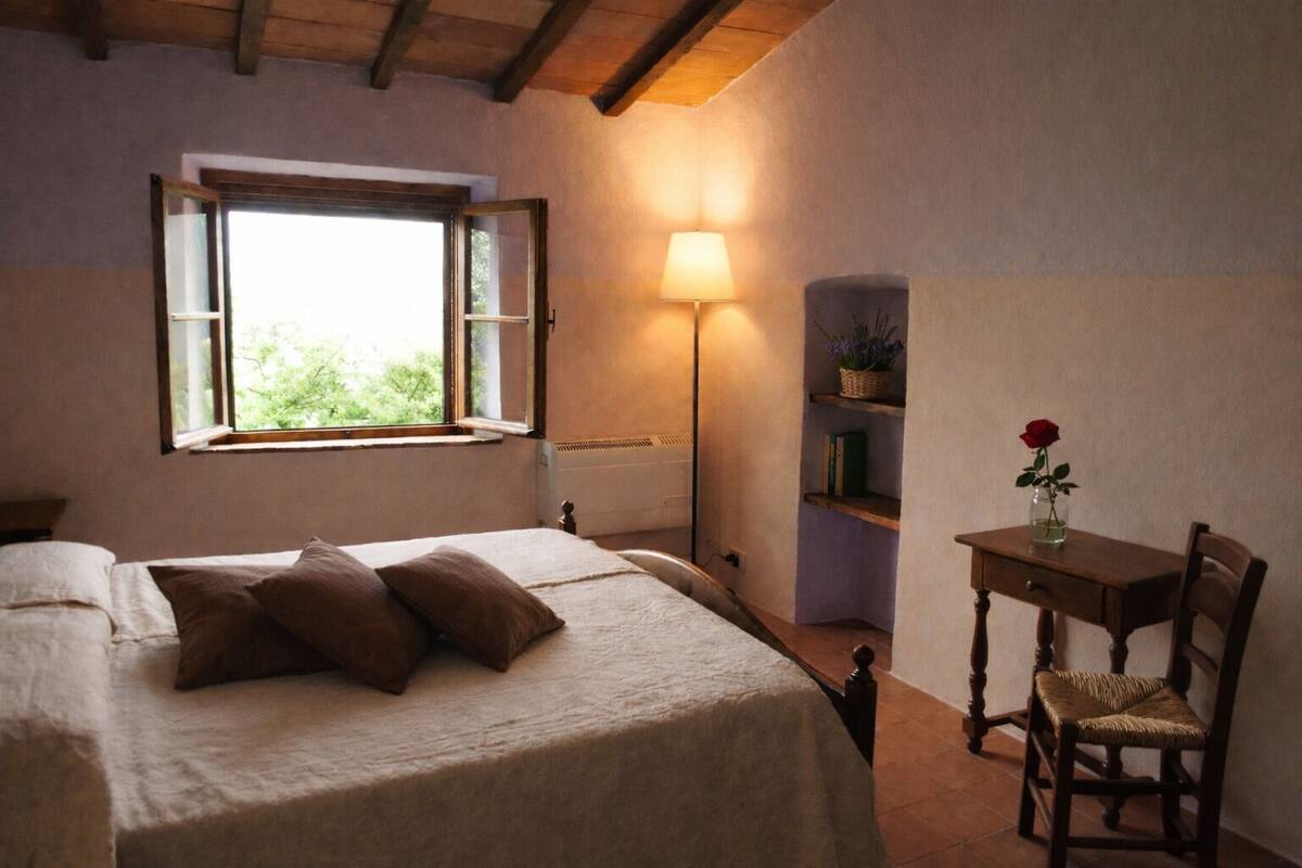 Un dormitorio con cama, escritorio, silla, lámpara y ventana con contraventanas abiertas.