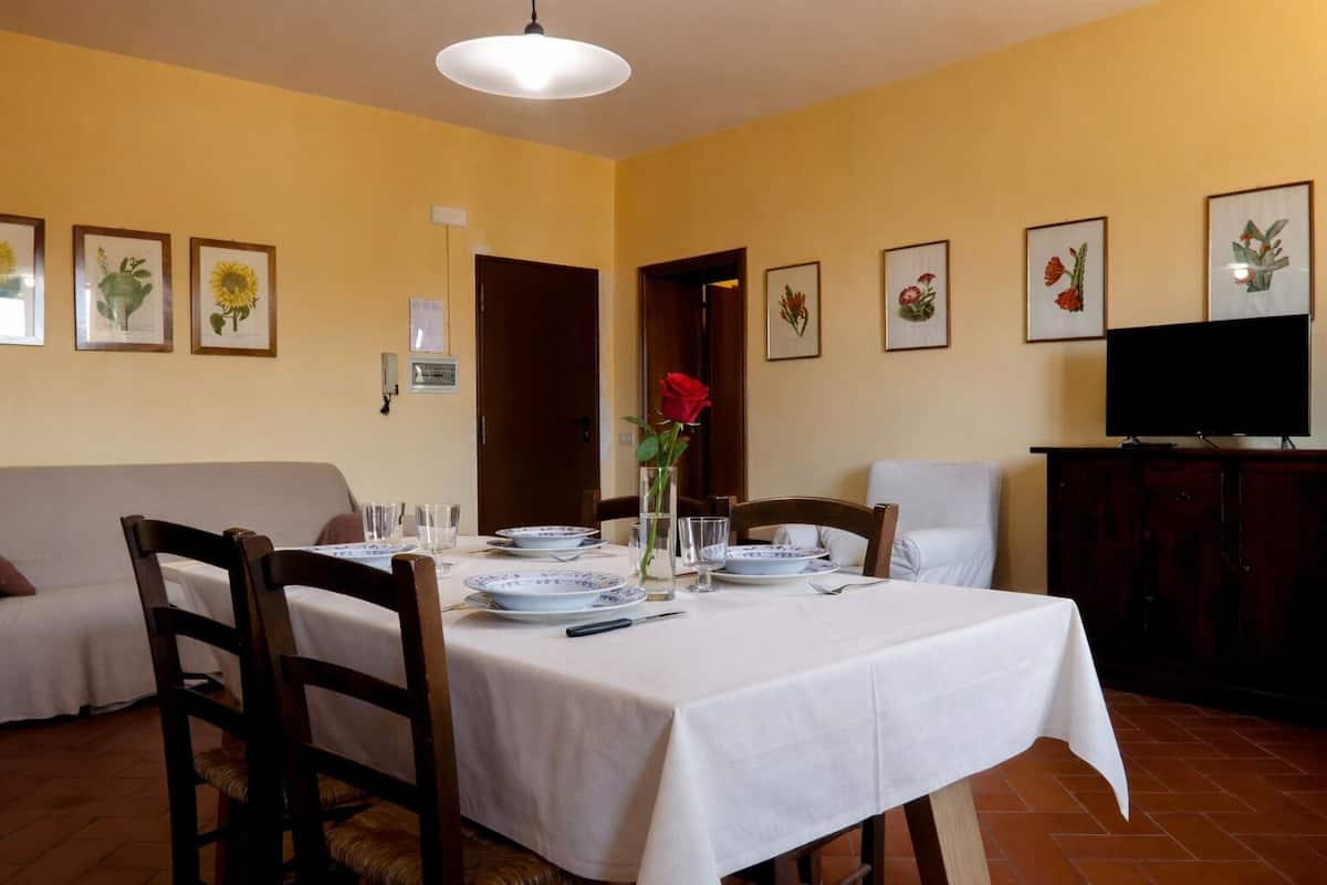 Comedor con mesa dispuesta para cuatro, sillas, un jarrón con una rosa roja e impresiones botánicas enmarcadas en las paredes.
