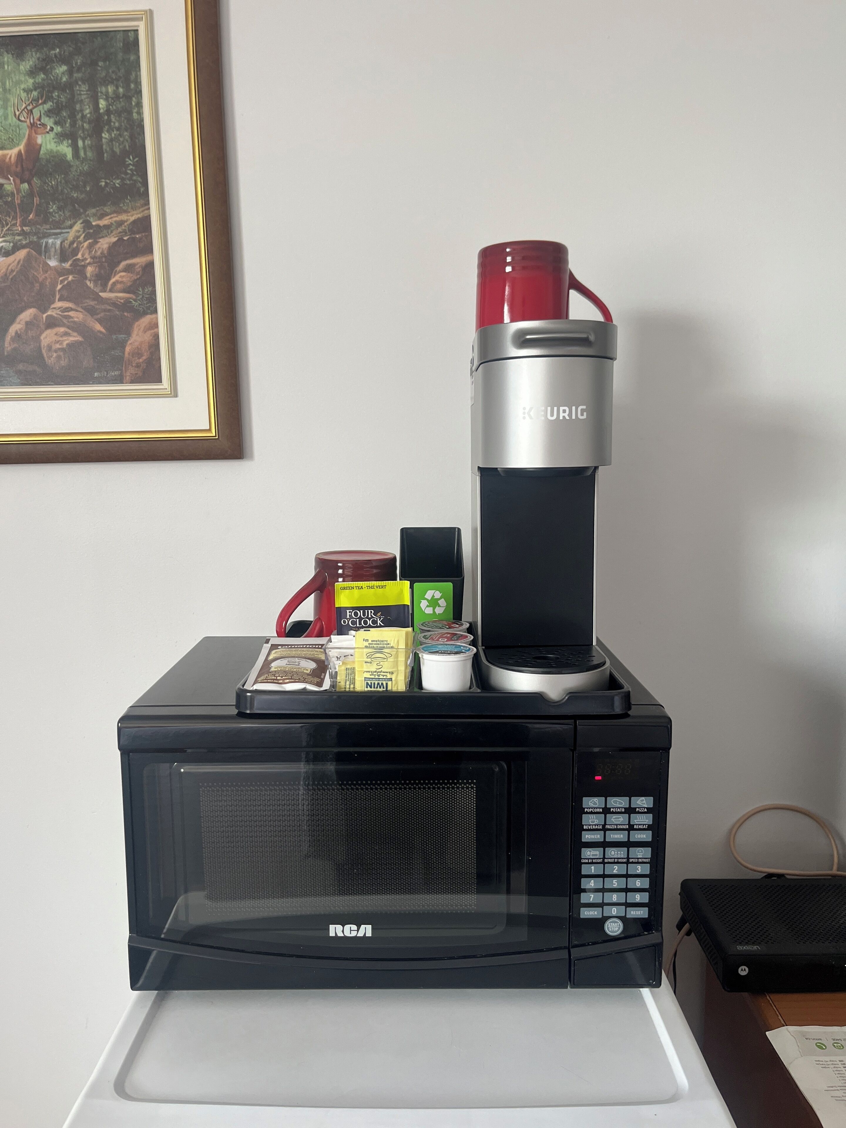 A microwave with a Keurig coffee maker on top, containing a red mug, a box of coffee pods, and a carton of milk.