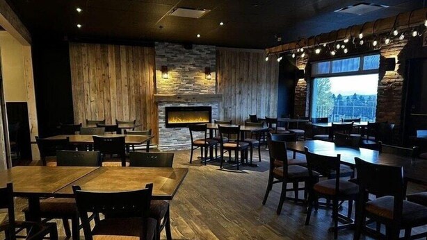 A restaurant with wooden tables and chairs, a stone fireplace, and a large window with a city view. - Hotel Motel Le Château (Lac-Megantic)