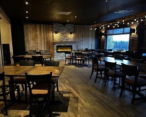 A restaurant with wooden tables and chairs, a stone fireplace, and a large window with a city view. - Hotel Motel Le Château (Lac-Megantic)