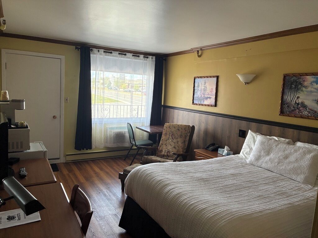 A hotel room with a bed, a desk with a lamp, a chair, a window with curtains, and two wall-mounted lamps.