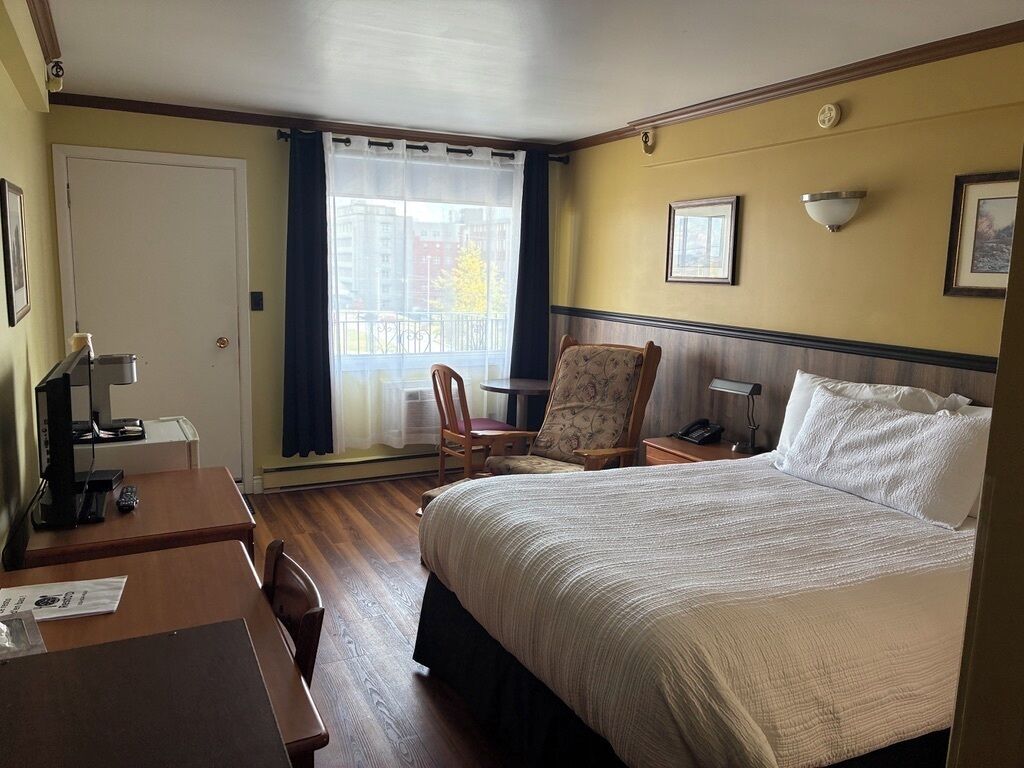 Ein Hotelzimmer mit einem großen Bett, einem Schreibtisch mit Computer, einem Stuhl, einem kleinen Tisch und einem Fenster mit Vorhängen.