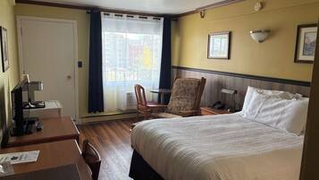 Ein Hotelzimmer mit einem großen Bett, einem Schreibtisch mit Computer, einem Stuhl, einem kleinen Tisch und einem Fenster mit Vorhängen.