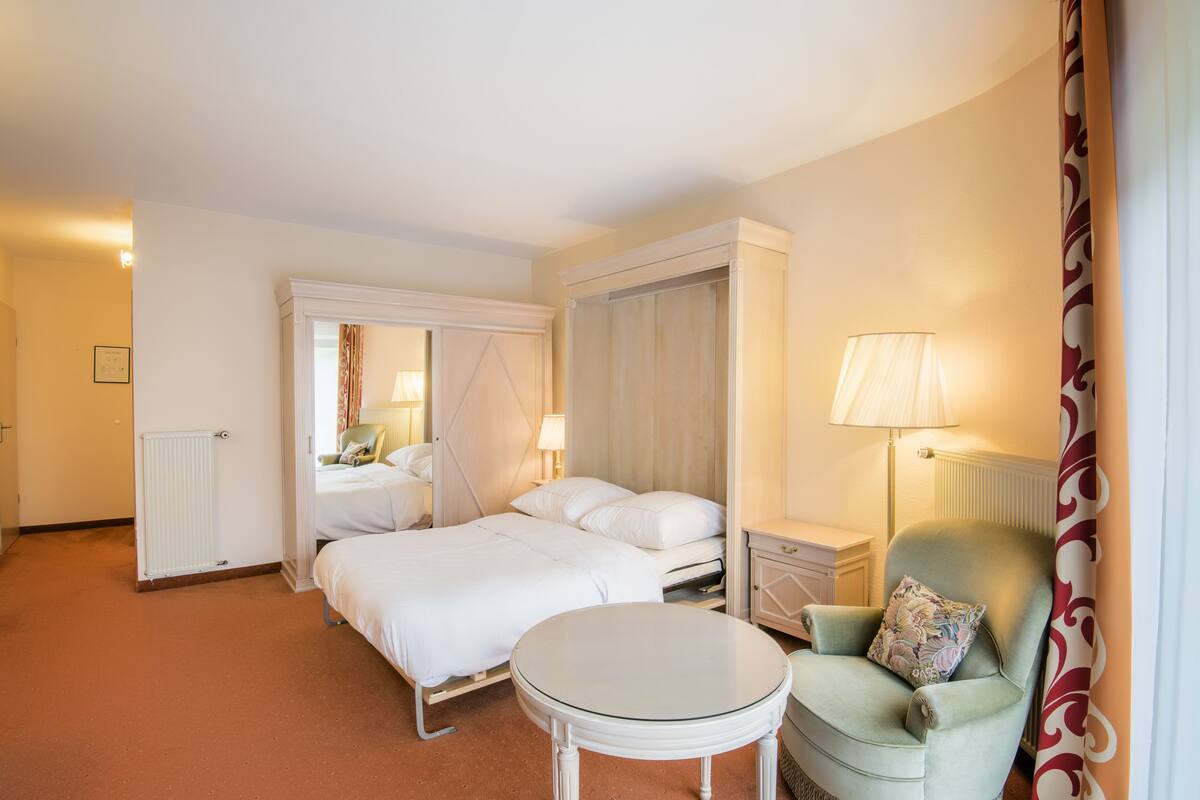 A hotel room with a four-poster bed, a round table, a chair, and a nightstand with a lamp.