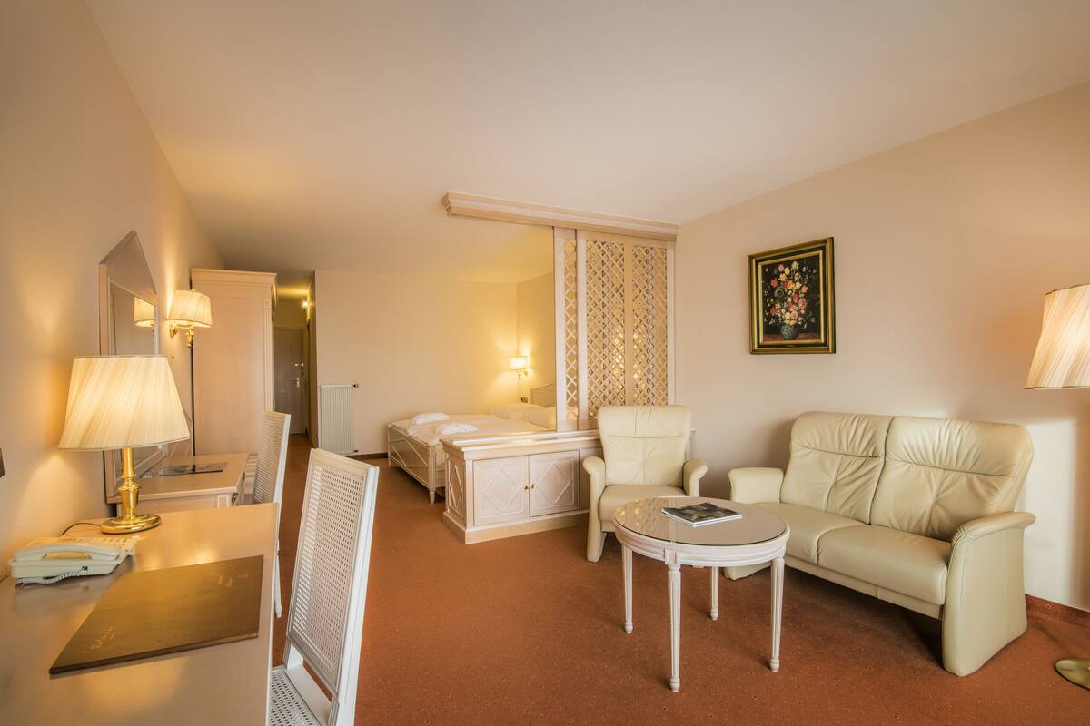 A hotel room with a bed, two chairs, a small table, a lamp, a painting, and a radiator.