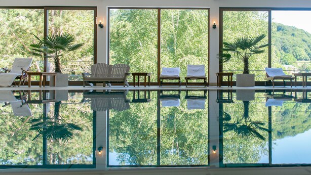 A pool area with chairs and tables, surrounded by large windows offering views of a forested hillside. - Hôtel Eden au Lac (Echternach)