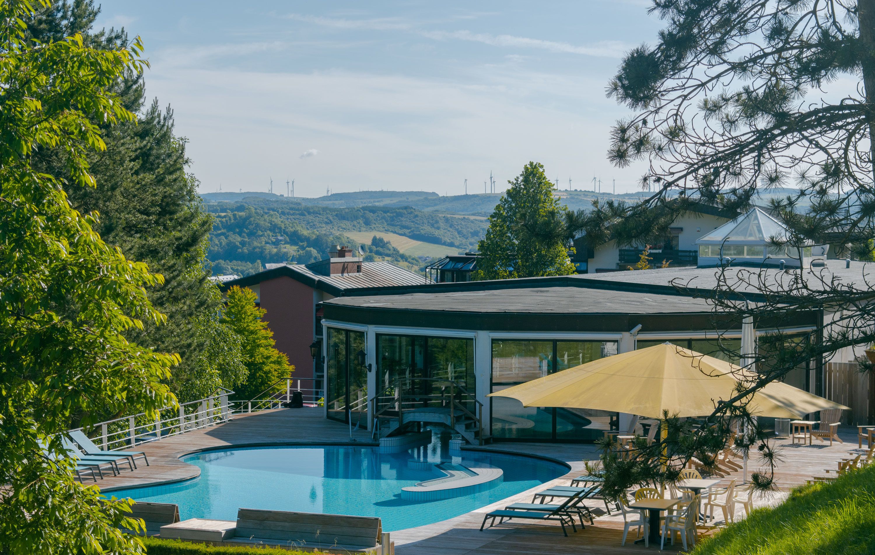 A swimming pool with lounge chairs and umbrellas, surrounded by trees and a building with a glass enclosure.