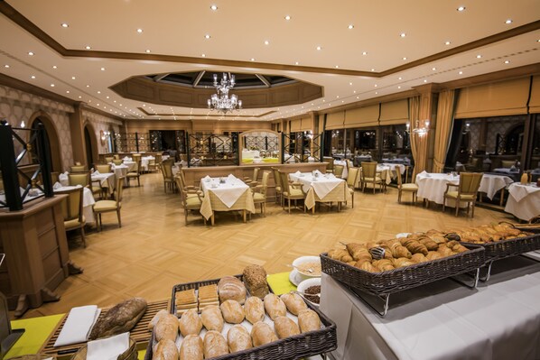 Amplia sala de comedor de hotel con varias mesas y sillas, un candelabro y una variedad de panes y pasteles exhibidos.