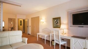 A hotel room with a white sofa, a round table, a desk with a chair, a television, and a painting on the wall.