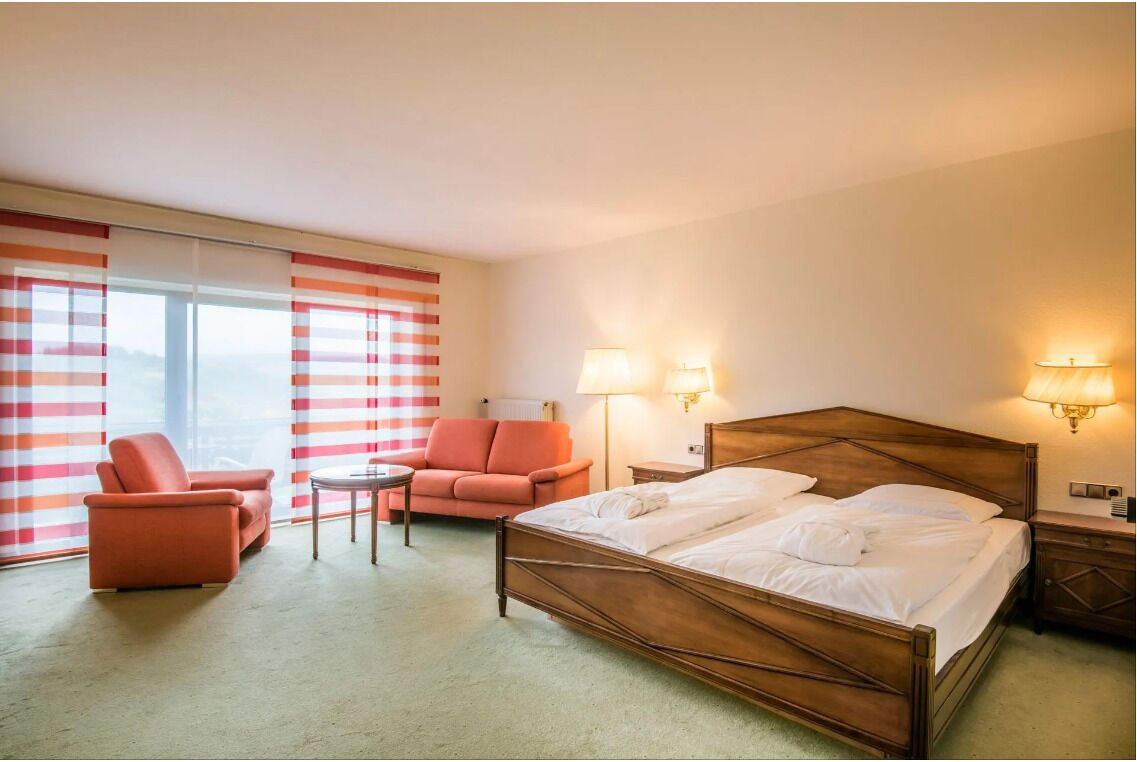 A hotel room with a bed, two chairs, a small table, and two bedside lamps.