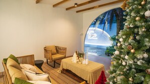 A cozy room with a wooden ceiling, a Christmas tree, a wicker sofa, and a view of the sea through a large window.