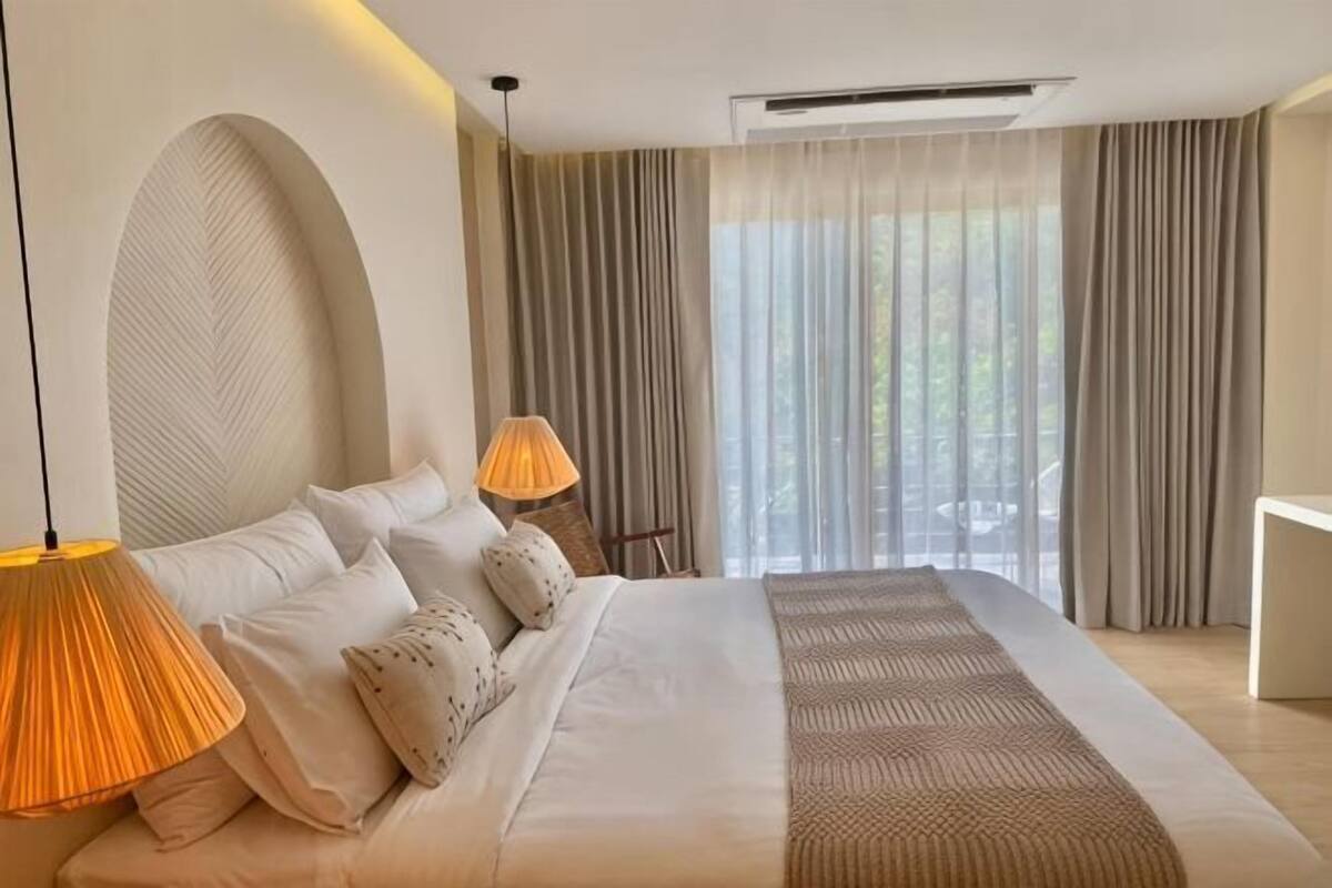 A modern hotel room with a large bed, two bedside lamps, a view of greenery through the window, and a ceiling vent.