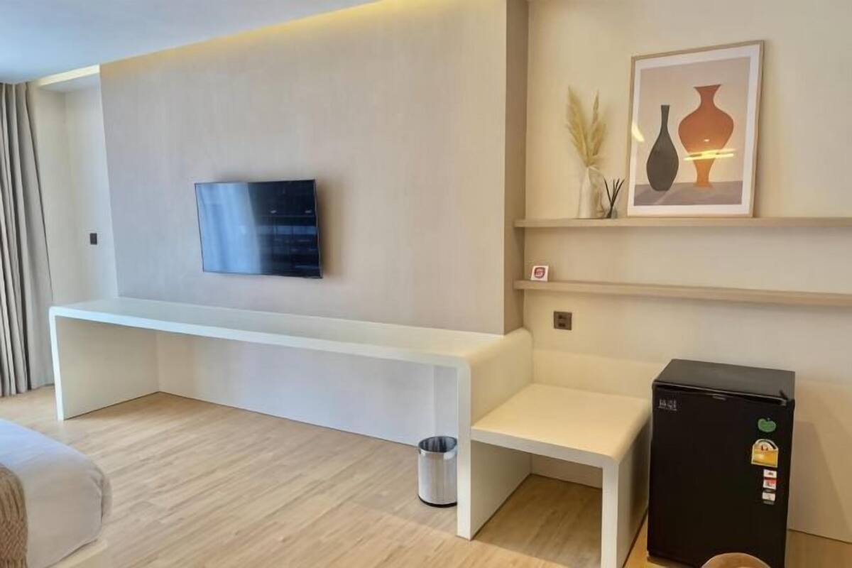A modern hotel room with a flat-screen TV mounted on the wall, a white bench, a small trash bin, and a black minibar.