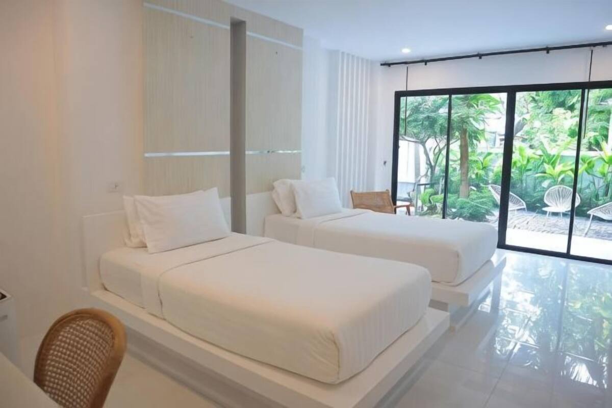 A modern hotel room with two beds, a large window with a view of greenery, and a bright, clean interior.