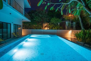 Una piscina con piso de baldosas, rodeada por un edificio blanco con balcón y una palmera.