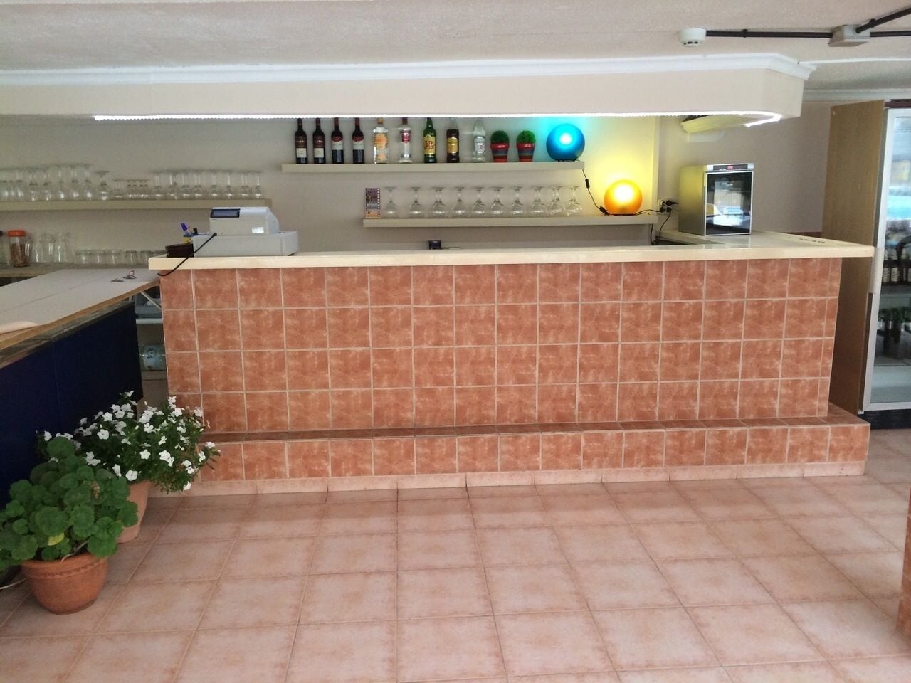 Een receptie met een betegelde balie, diverse flessen en glazen op planken, een koffieapparaat en potplanten.