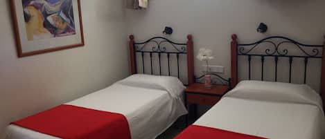 Una habitación con dos camas individuales con colchas rojas, una mesita de noche con un jarrón de flores, un cuadro abstracto enmarcado y una ventana con cortinas.