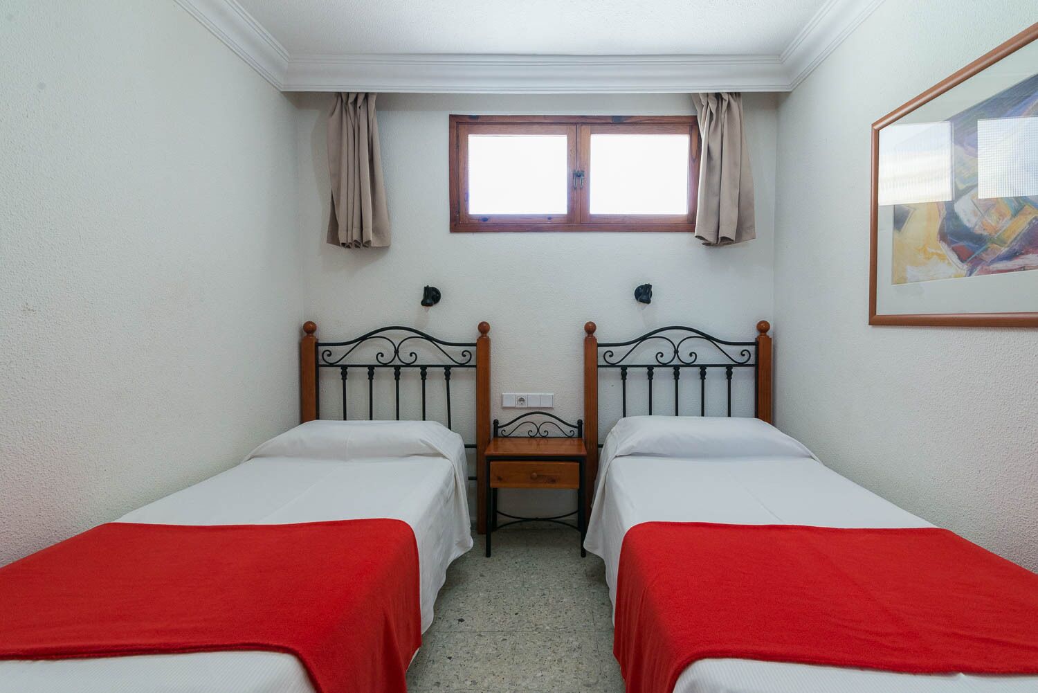 A small, clean room with two single beds, a wooden nightstand, a window with curtains, and a painting on the wall.