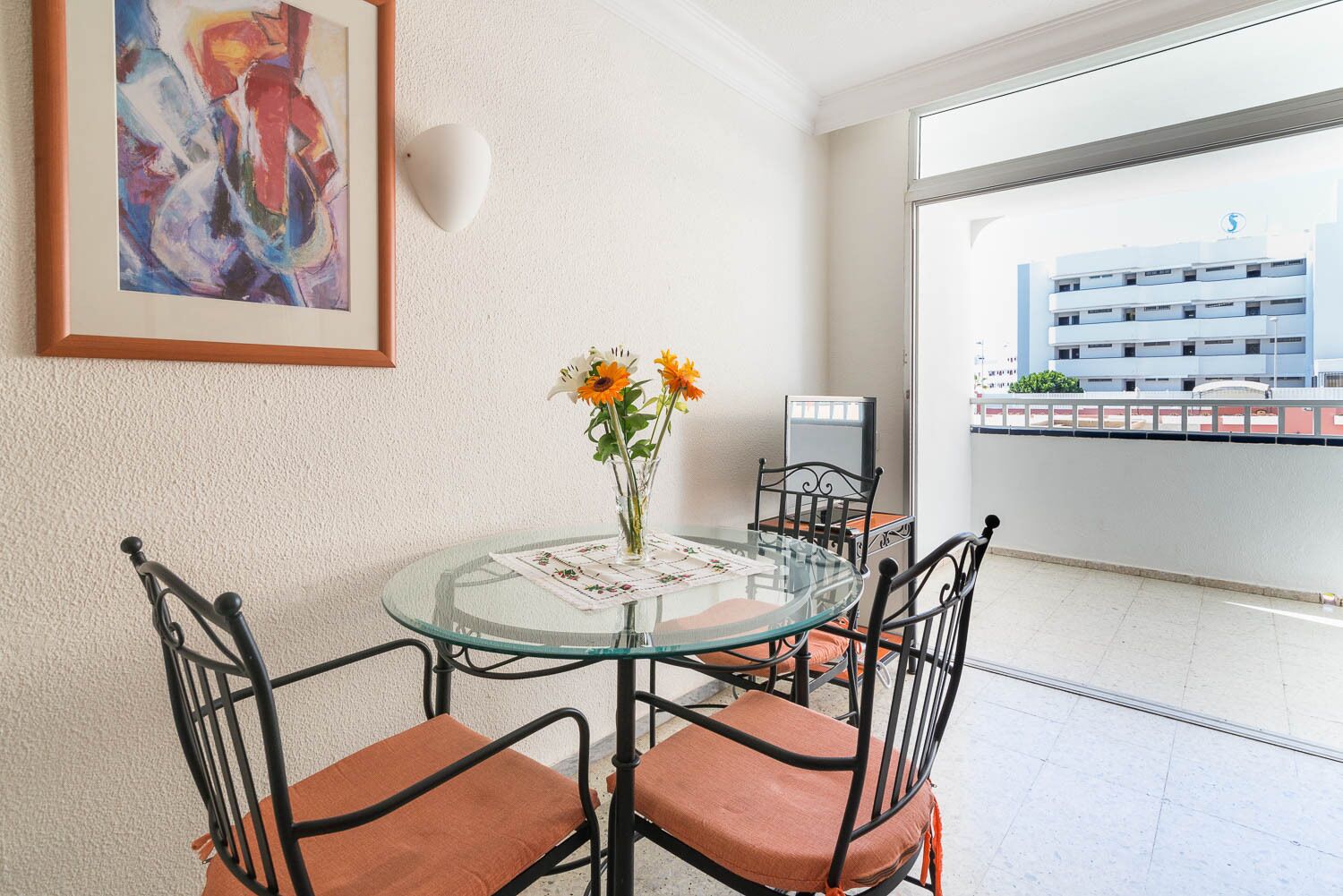 Un coin repas avec une table en verre, quatre chaises, un vase de fleurs et un tableau accroché au mur.