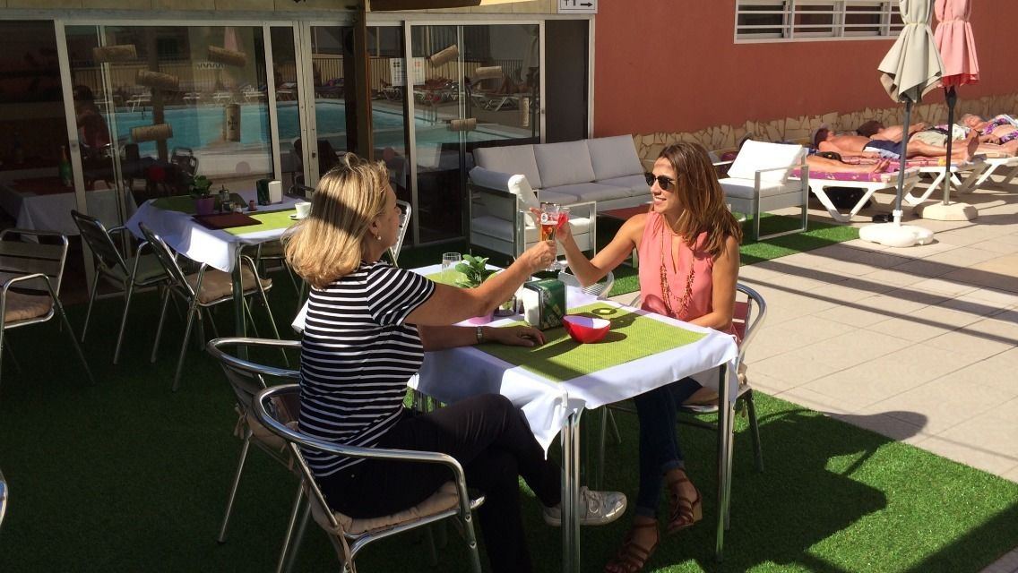Dos personas sentadas en una mesa al aire libre con vista a una piscina y sillones de descanso al fondo.