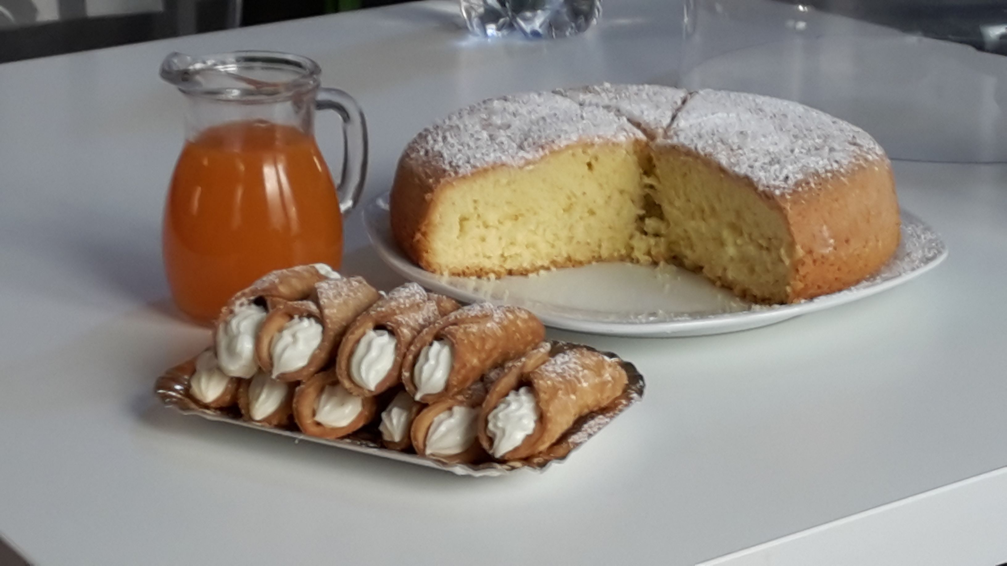 A cake with a slice missing, a pitcher of tea, and a tray of cream-filled pastries on a white table.