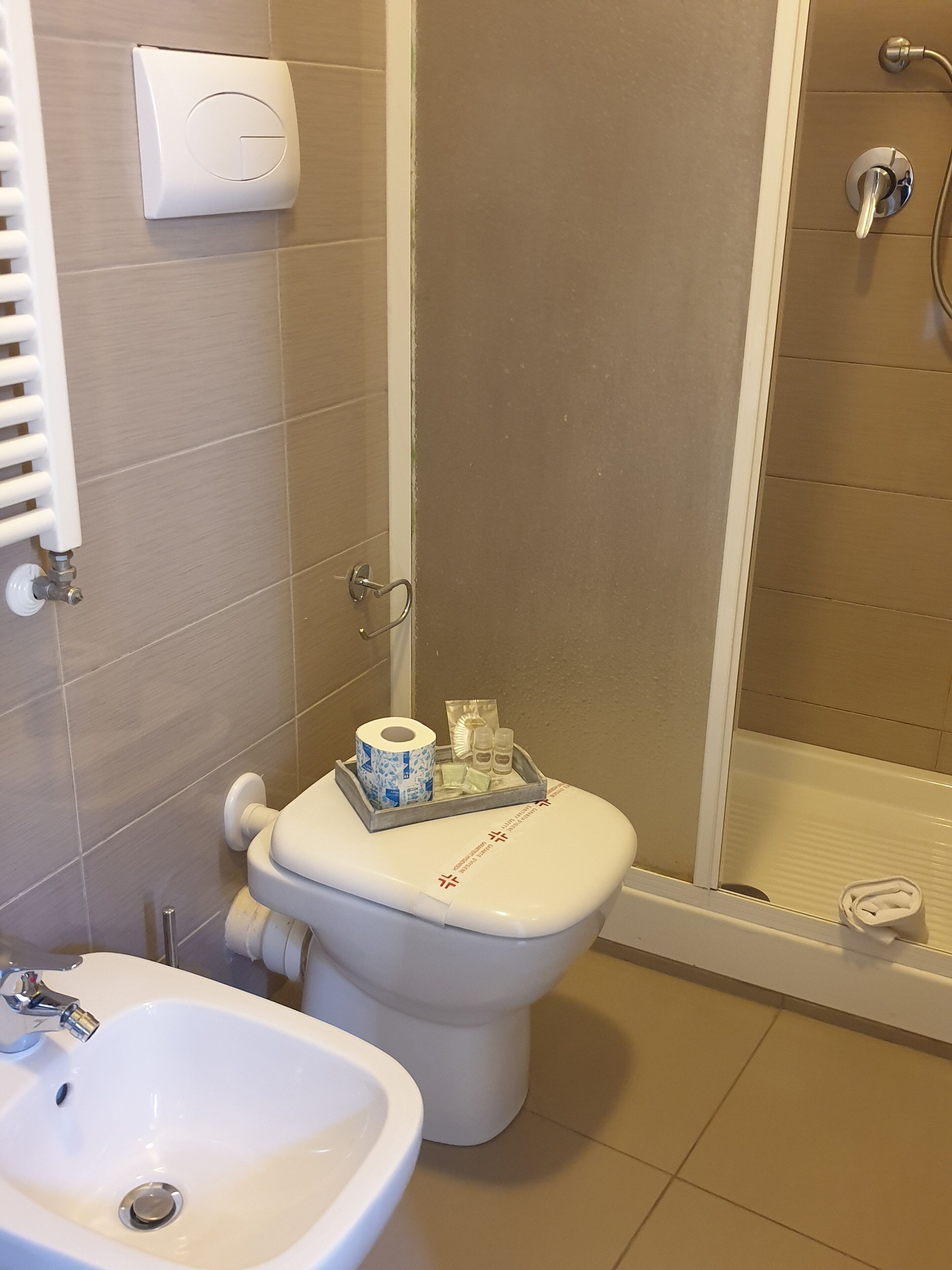 A bathroom with a toilet, bidet, and sink. There is a towel dispenser and a shower area visible.