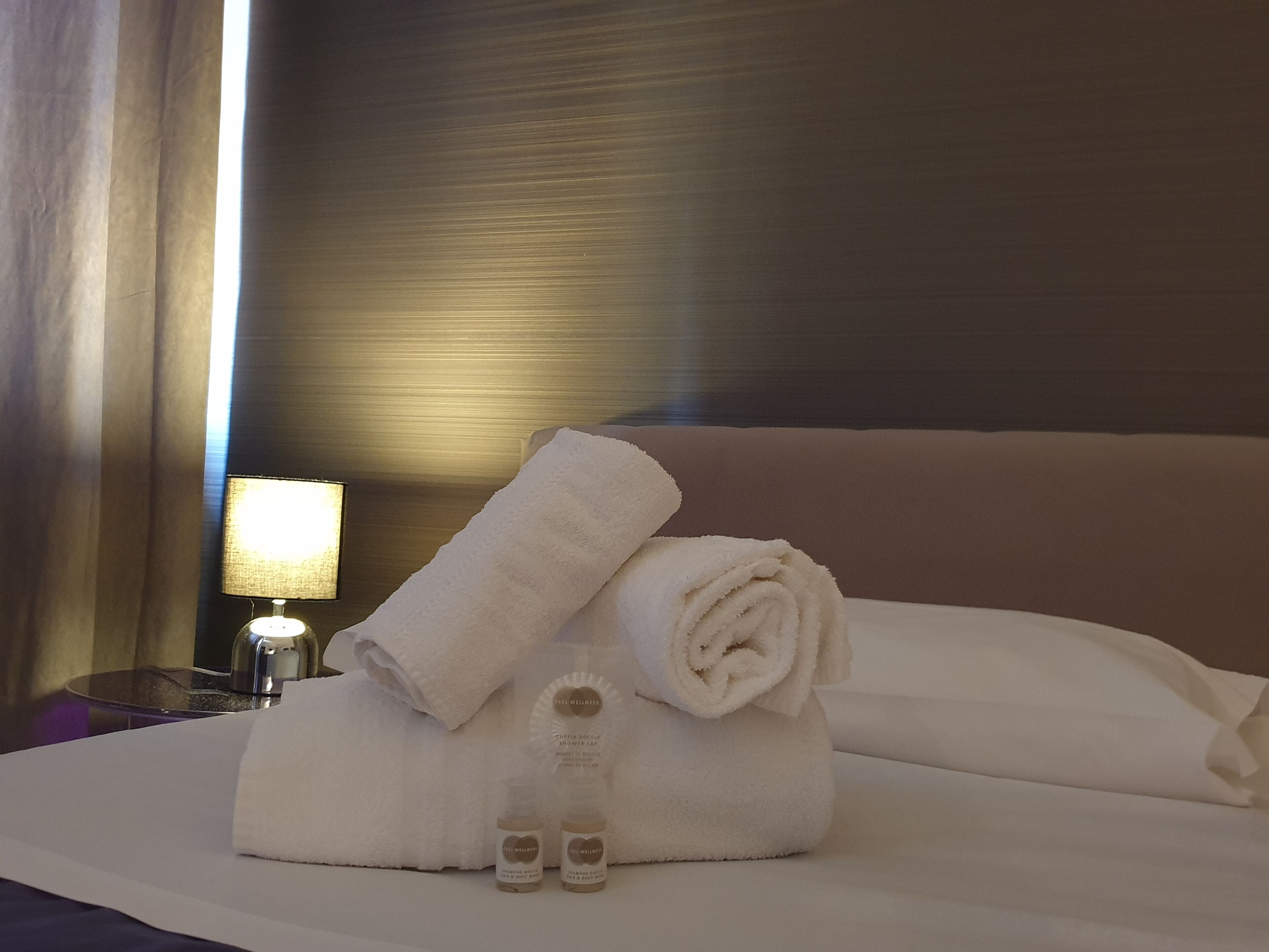 A neatly made bed with a folded white towel, two bottles of lotion, and a lamp on the side.
