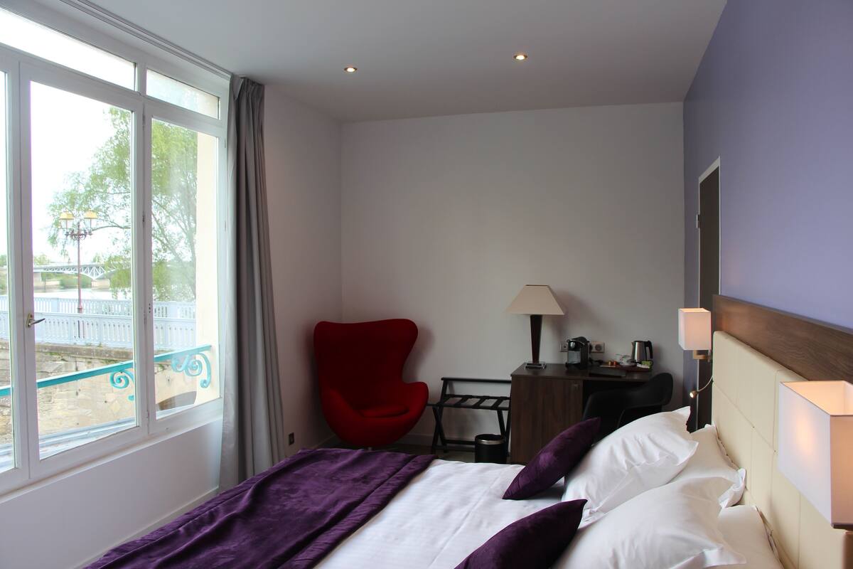 A hotel room with a bed, a red chair, a desk with a lamp, and a view of the outside.